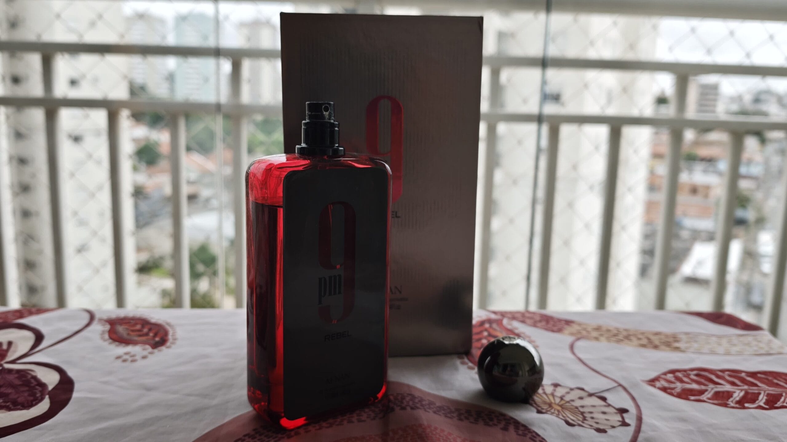 Afnan 9PM Rebel with the cap off and the atomizer showing, the silver box behind it, on a balcony table