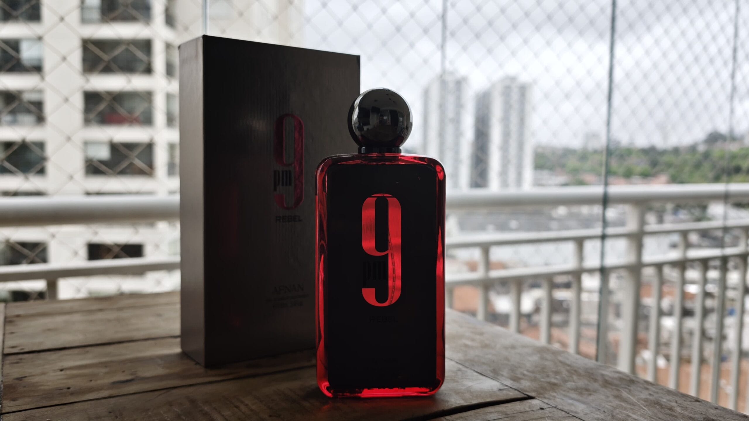 Afnan 9PM Rebel with its chrome cap on, the box behind it, on a wooden table with the city in the background